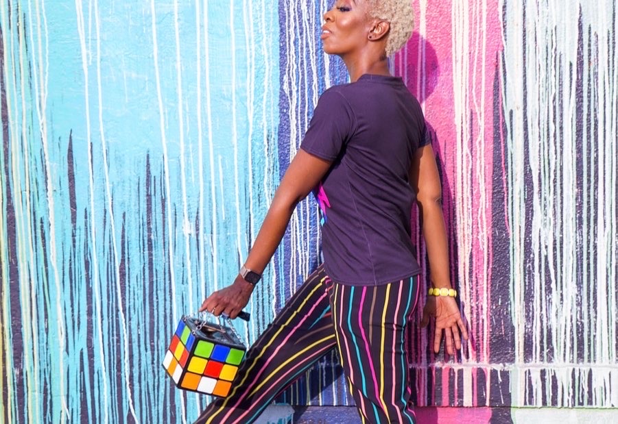Ebony Noir leaning against a brightly painted color wall, holding a Rubik's cube handbag